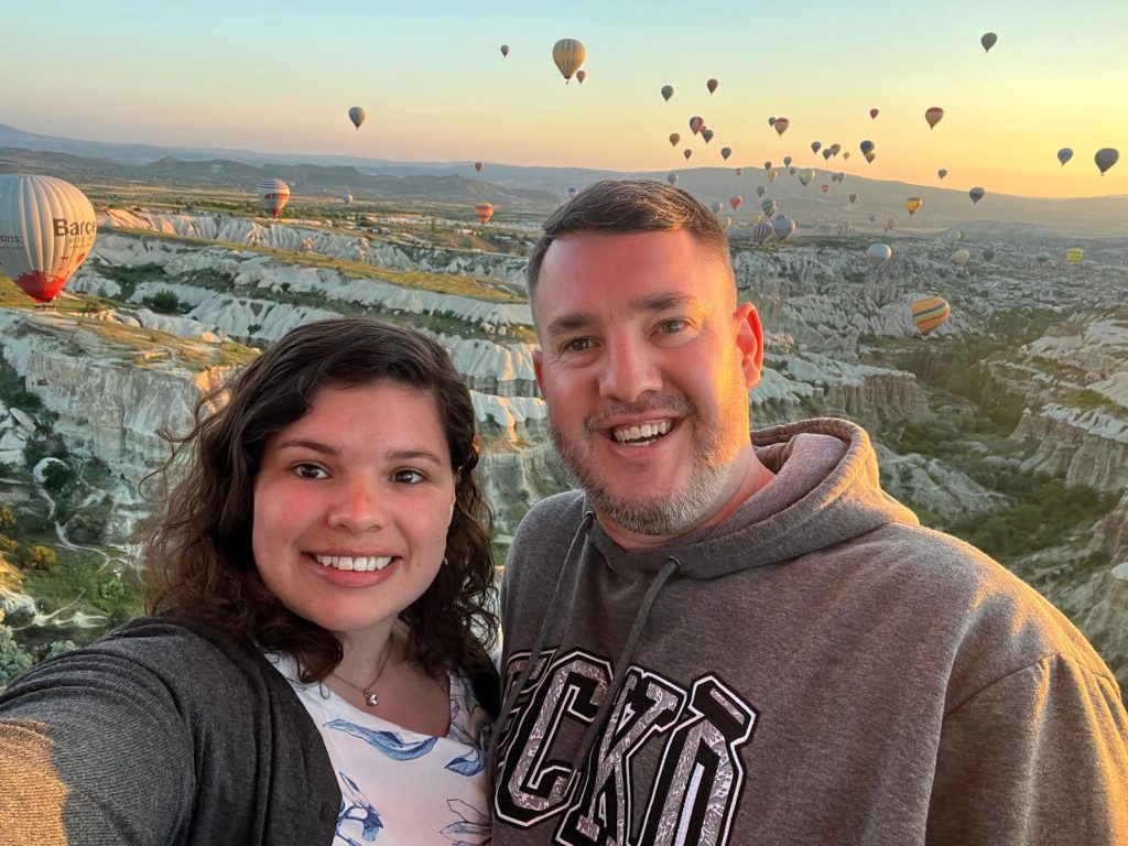 couple standing in front of a background where hundreds of hot air balloons fill the sky during sunrise in Cappadocia, Türkiye.