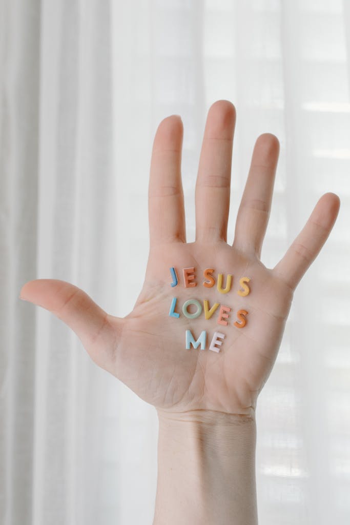 An open hand with colorful letters reading 'Jesus Loves Me' against a soft curtain backdrop.