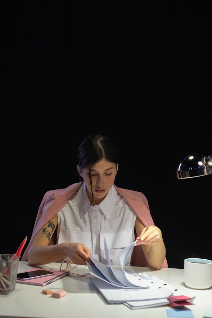 Woman in pink blazer reviewing papers late at night in dimly lit office.