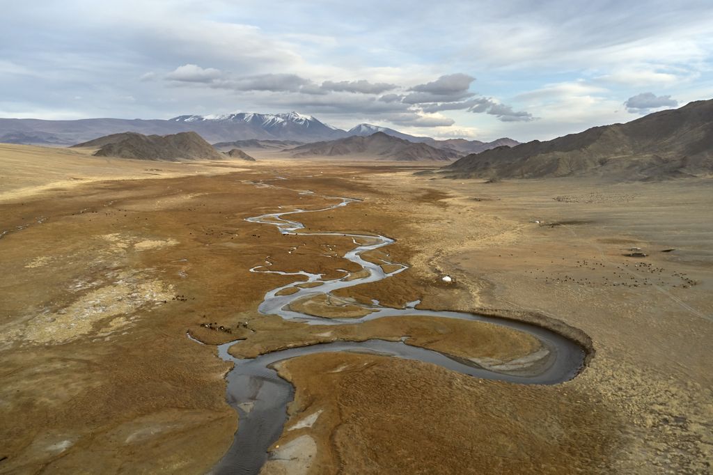 Aerial view of scenic vast dry semidesert terrain surrounded with mountains and narrow curvy river against cloudy sky