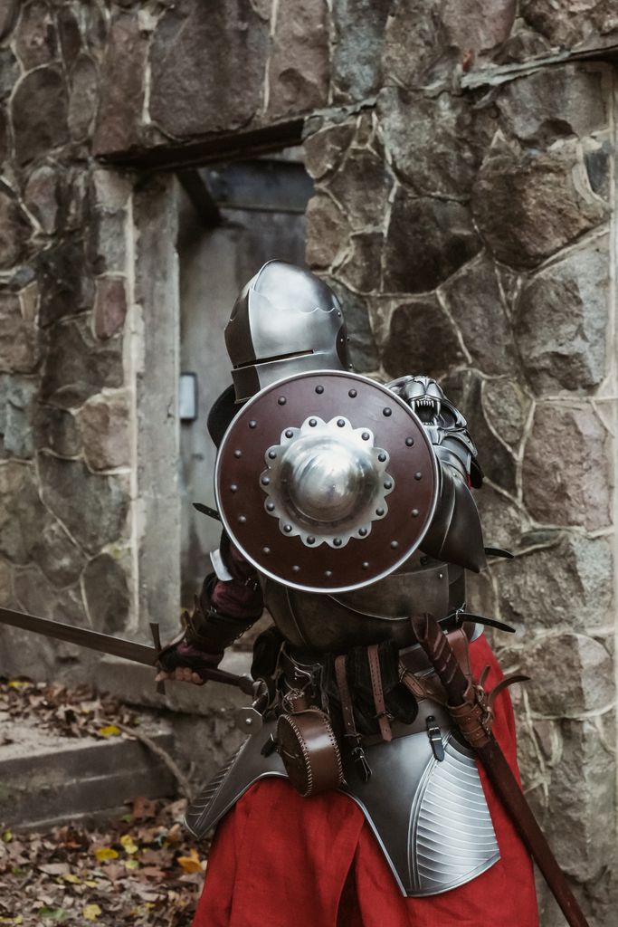Back view of a medieval knight in full armor standing by a stone wall, carrying the shield of faith that will protect him from the flaming arrows of satan.