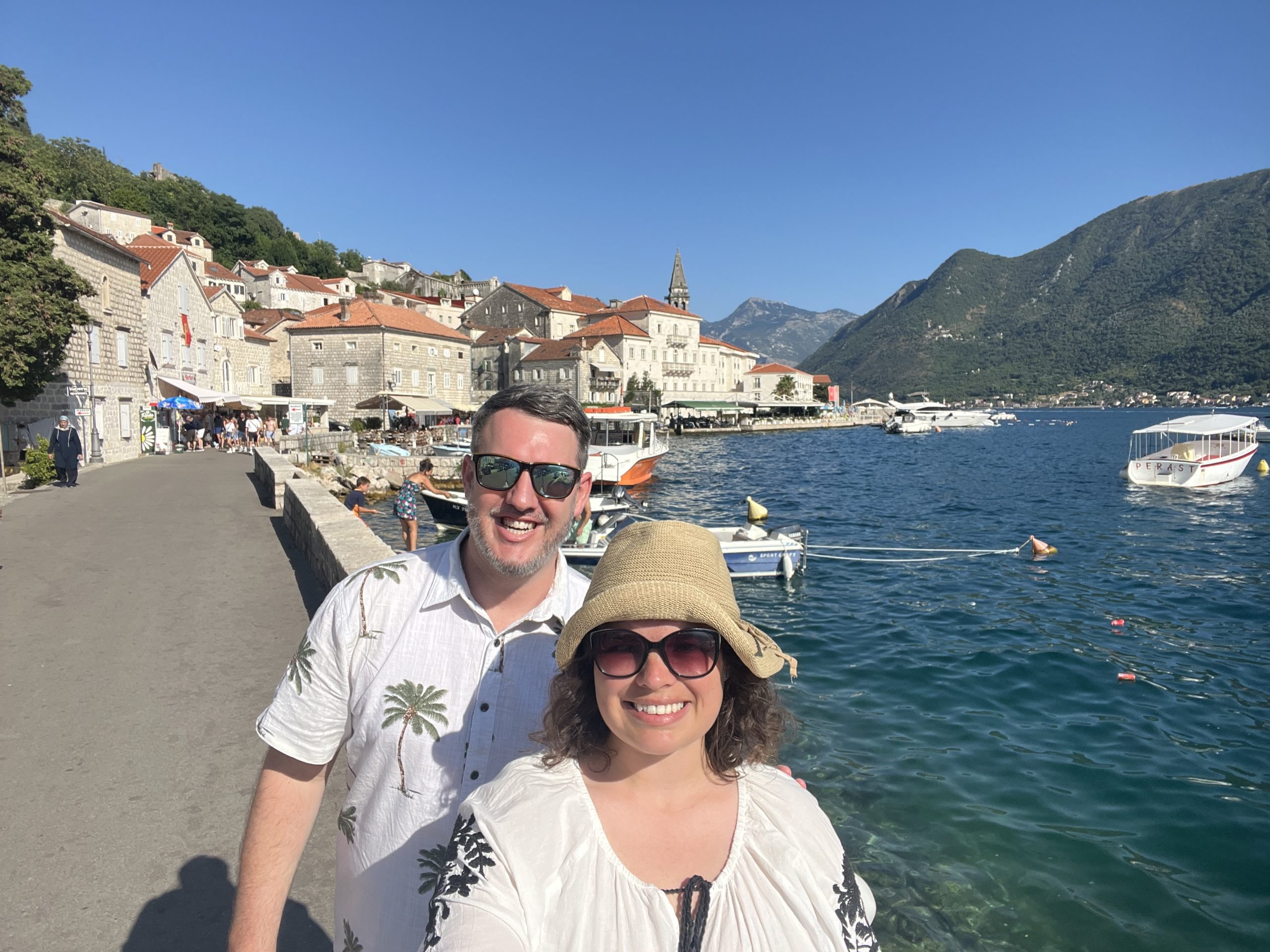Couple standing in front of historic buildings with red roofs in Perast, Montenegro with the blue waters of the Adriatic Sea in the background.