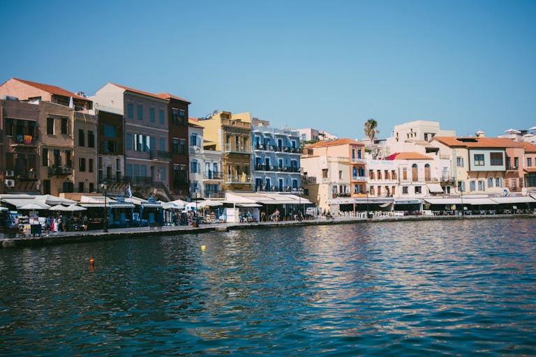 Colorful historic buildings line the waterfront of Chania's Venetian harbor under a clear blue sky.