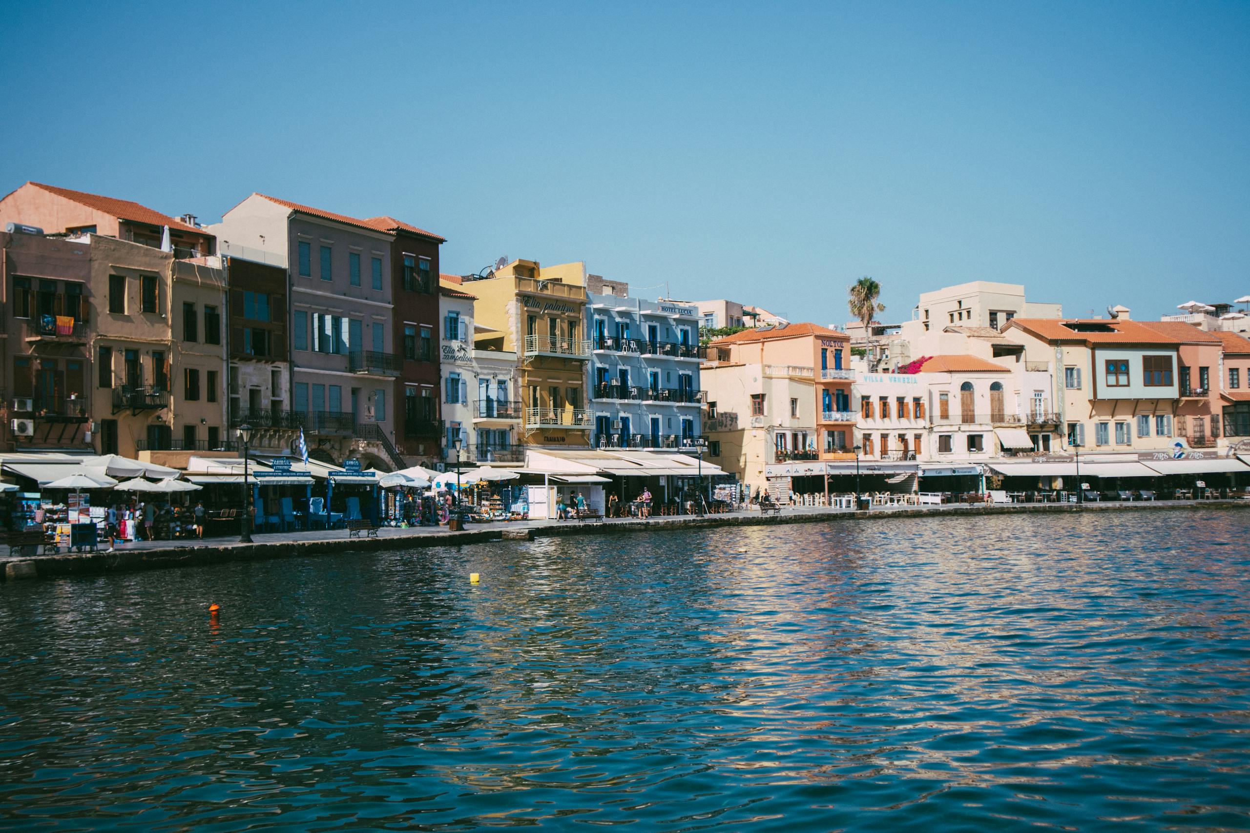 Colorful historic buildings line the waterfront of Chania's Venetian harbor under a clear blue sky.