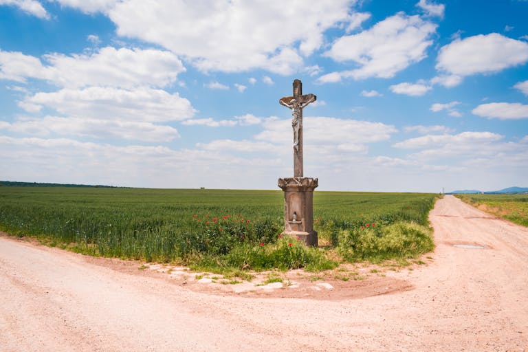 A solitary cross at a rural crossroads under a blue sky with white clouds.