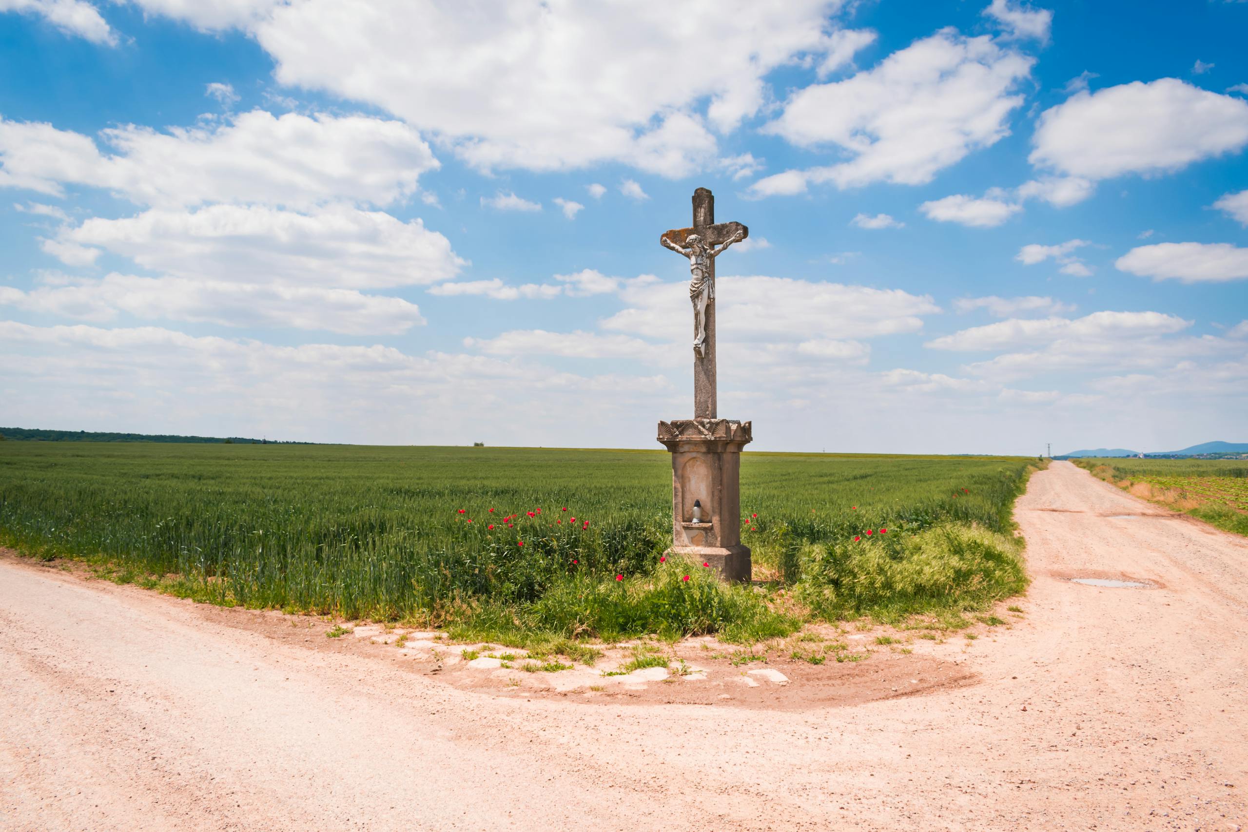 A solitary cross at a rural crossroads under a blue sky with white clouds.