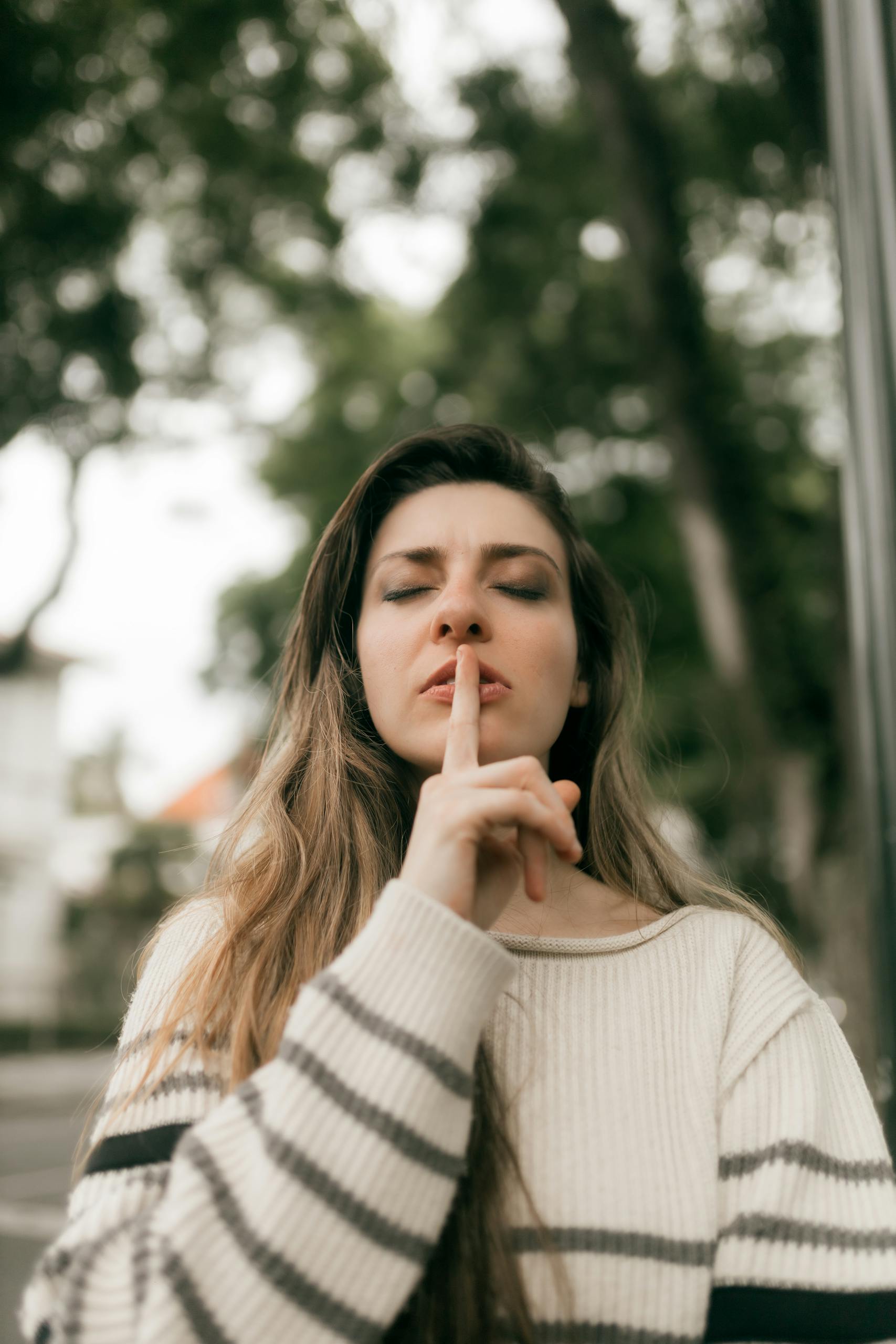 A woman outdoors, wearing a striped sweater, gestures for silence in a serene setting.